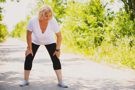 Sporty blonde woman resting after jogging in the park in the morning wearing smart watch and keeping her hands on her knees.の写真素材