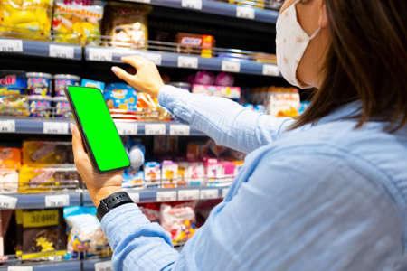 Cropped photo of a young woman with face mask using mobile phone and buying groceries in the supermarket during virus pandemic.の写真素材