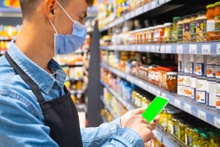 Side view photo of a store worker man in a medical mask wearing apron black color holding the smartphone and doing the inventory of the products on the shelves.の写真素材