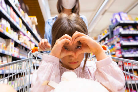 Close up photo of a girl making heart sign with her hands and having serious look, young woman pushing the cart wearing medical mask being in the store.の写真素材