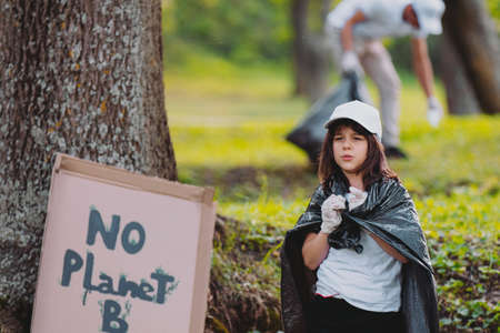 Photo of a brunette young girl volunteer worker standing with a garbage bag covering her shoulders outdoors with handmade cardboard banner attached to the tree, save planet.の写真素材