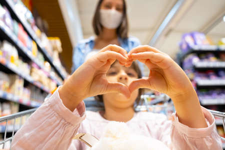 Pretty young girl making heart sign with her hands while sitting in the shopping cart and her mother standing behind her in a medical mask, family shopping concept.の写真素材