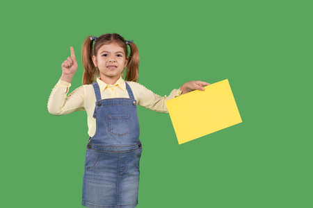 Little schoolgirl shows thumbs up at green background holding the yellow paper in her hand and pointing with her finger up, paying attention.の写真素材