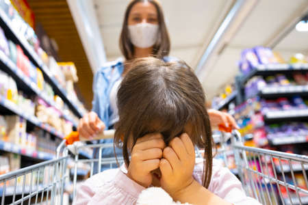 School aged girl covers her face with the hands sitting in the shopping cart and her young mother wearing medical mask pushing the cart passing among the shelves in the store.の写真素材