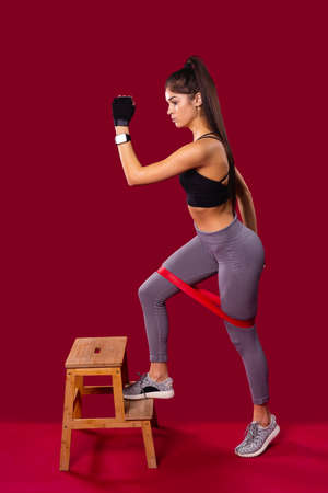 The frontal side pose of a very beautiful slim girl practicing fitness exercises using elastic band on her legs lifted her one leg to the step of the chair and raised one hand up.の写真素材