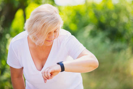 Close up photo of a blonde sporty woman wearing white t-shirt looking at the smart watch on her hand and checking the pulse after jogging.の写真素材