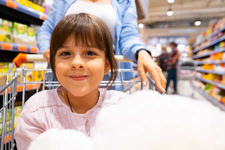 Pretty nice girl looking into the camera while sitting in the shopping cart and her young mother pushing the cart being in the store buying products to bring them home.の写真素材