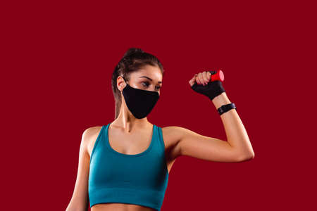 Fitness girl with a textile mask posing in gym in blue top holding one red dumbbell in her hand raised it bending her arm in the log.の写真素材