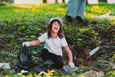 Close up photo of a laughing young girl sitting down on the ground in white gloves holding a garbage bag and picking up plastic rubbish.の写真素材