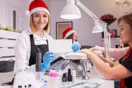 Young woman professional manicure specialist wearing a santa hat and protective gloves holding white voucher for the client opposite for new year holiday.の写真素材