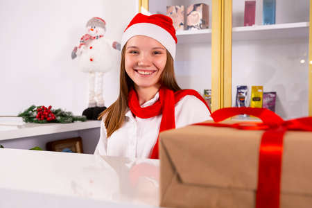 Smiling office manager sitting in the workplace wearing Santa hat and red scarf and present box with the red ribbon lies on the table.の写真素材