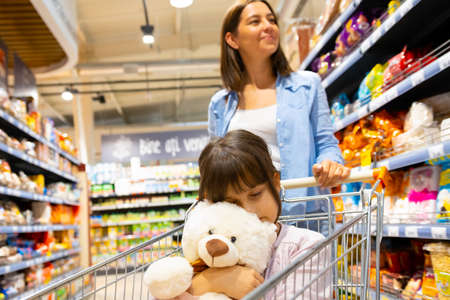 Girl in the shopping cart holding the toy and sleeping, while her mother looking for the products to buy in the store.の写真素材