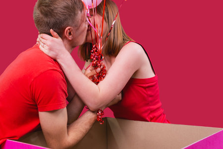 Young beautiful couple in red t-shirts are kissing on the red background, blonde lady accepted marriage proposal from her boyfriend holding balloons.の写真素材