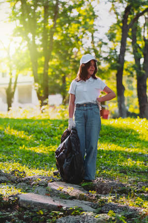 Full-length photo of a smart young girl standing on the stones in the park keep her hand on the waist looking away and holding the garbage bag in the other hand.の写真素材