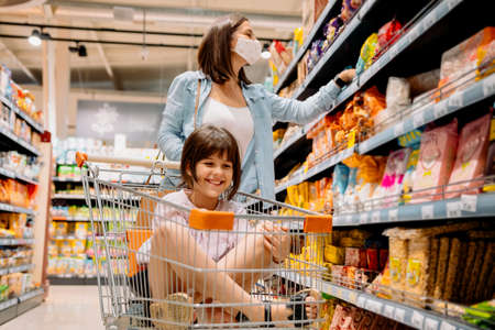 Close up photo of two female persons in the store, mother in the mask looking for the goods on the shelf and her daughter sitting in the grocery cart with smiling face.の写真素材