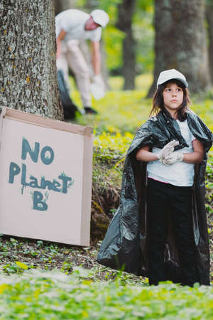 Full-length photo of a pensive girl with protective gloves and garbage bag on her shoulders standing near a cardboard banner with the inscription, a man over the background outdoors, love the nature.の写真素材