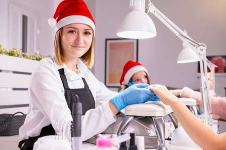 Professional young woman manicurist in a Santa hat and protective blue gloves holding the client hands an looking at the camera.の写真素材