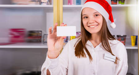 Beautiful young girl office manager in the company smiling wearing the Santa cap having the badge on her white shirt and holding in her hand holiday voucher, copy space.の写真素材
