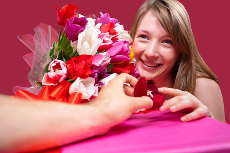 Close up photo of a beautiful joyful blonde lady holding a tulips colored bouquet and accepting the marriage proposal from her boyfriend, he holding a small box with a ring in it.の写真素材