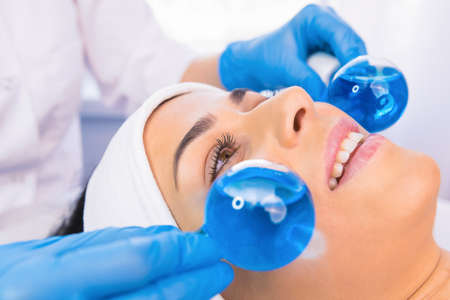 Close up portrait of beautiful woman with towel on head undergoes massaging face procedure by a cosmetologist using blue ball roller in a beauty salon.の写真素材