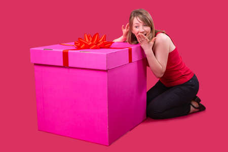 Close up photo of a young pretty lady in red t-shirt and black jeans sitting on the floor near the big present box of pink color and covers her mouth with her hand having surprised face expression.の写真素材