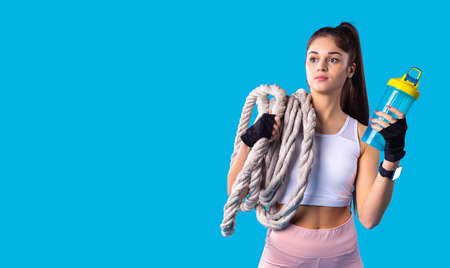 Indoor shot of attractive young active brunette young girl with ponytail hairstyle holding bottle of water and keeping the fitness rope on her shoulder.の写真素材
