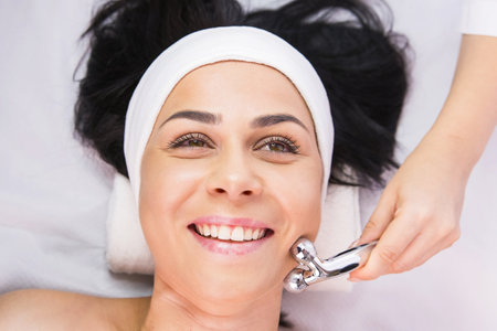 Close up photo of a happy client woman in a beauty salon, professional cosmetologist making her relaxing face massage using cosmetic roller.の写真素材