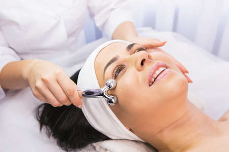 Relaxed young woman undergoes the rejuvenation facial massage procedure in a beauty salon by a cosmetologist using face roller.の写真素材
