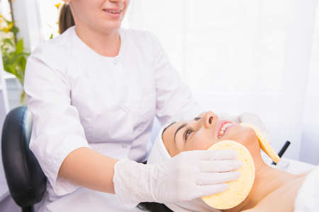 Professional cosmetologist making cleansing procedure for moisturizing the face with two sponges to a woman in a beauty salon.の写真素材