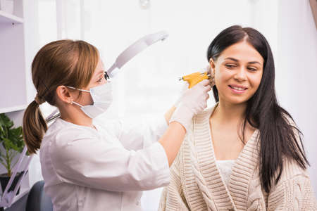 Specialist in medical mask and gloves pierces the clients ears with a piercing gun in the cosmetologist cabinet.の写真素材