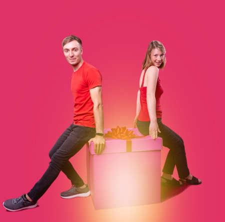 Beautiful young couple in red clothes and black jeans sitting on the present box with their backs to each other and they are happy.の写真素材