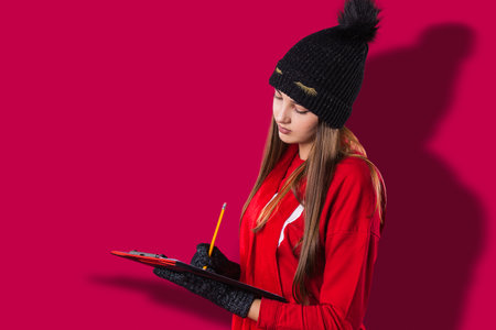Side view photo of a pretty girl dressed in red clothes, black hat and gloves holding the document in her hands and writing with a pencil.の写真素材