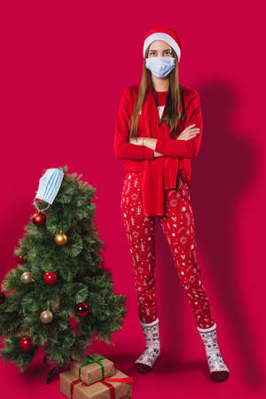 Full-length photo of a young woman dressed in red pajamas having a Santa cap on her head wearing medical mask standing with cross hands near the Christmas tree with a medical mask on the top.の写真素材