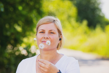 Close up photo of a happy sporty blonde woman outdoors wearing smart watch and earphones holding the dandelion flower in her hand.の写真素材