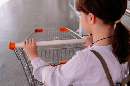 Back view photo of a nice girl pushing the empty grocery cart outside the supermarket, wearing white blouse and backpack.の写真素材