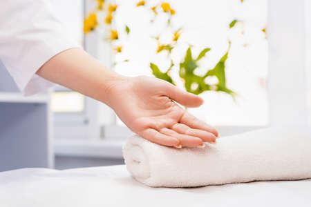 Close up photo of a cropped cosmetologist woman hand, female palm up on a folded white towel in a beauty salon, health and clean concept. Yellow natural flowers. Ready for massageの写真素材