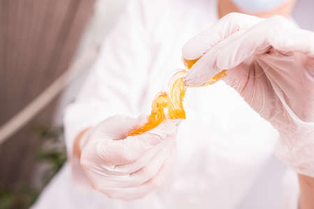 Selective focus. Close up photo of a cosmetologist woman hands with protective gloves holding honey wax shugaring paste in a beauty salon.の写真素材