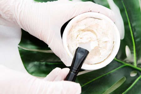 Photo of a specialist hands wearing medical gloves holding brush and professional cream peeling clay white mask in bowl, salon procedure, skin treatment.の写真素材