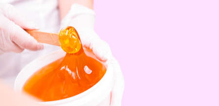 Banner- long format. Close up photo of a bowl with ,shugar paste, shugaring wax gold color hold by a professional specialist woman cosmetologist, skin care concept.の写真素材