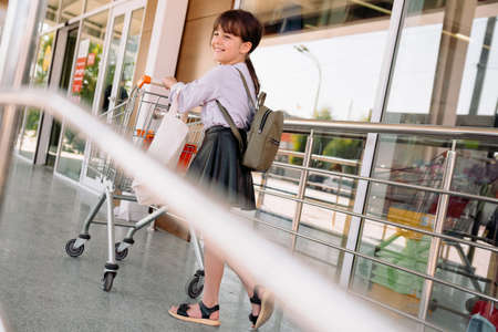 Joyful girl wearing modern clothes holding the bag with handles white color and backpack on the back pushes the grocery cart to enter the supermarket.の写真素材