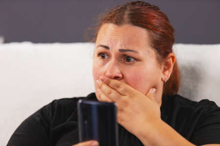 Close up photo of an worried woman in black t-shirt sitting on the white sofa holding the smart phone in her hand and watching bad news on tv covering her mouth with her hand.の写真素材