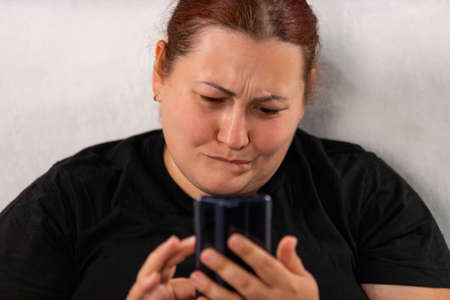 Close up photo of a woman with sad facial expression in black t-shirt holding the mobile phone in her hands and browsing bad news headlines.の写真素材