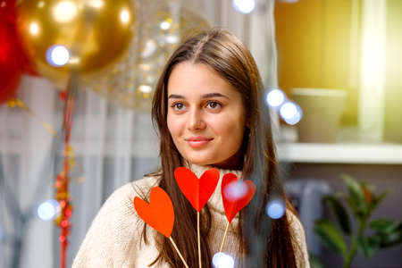 Close up photo of a happy young lady holding red heart shape flowers of paper on the new year party with balloons over background.の写真素材
