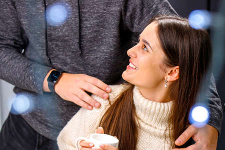 Happy smiling young lady in white warm sweater holding a cup of tea in her hands looking up at her boyfriend hugging her shoulders.の写真素材
