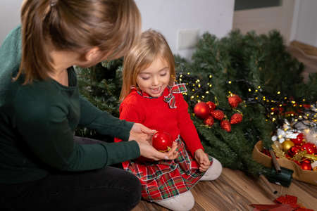 Happy little girl sitting on the floor with her mother with details of Christmas tree and smiling widely because her mother gave her a beautiful red decorative ball.の写真素材