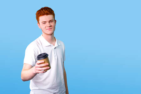 Young man with red hair wearing white t-shirt holding in his hand a cardboard cup of coffee, standing on the blue background.の写真素材