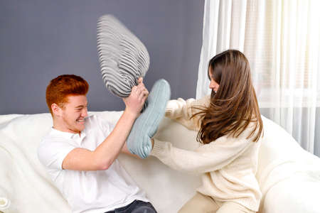 Young couple red hair man and pretty woman having fun, holding pillows in their hands and fighting sitting in the sofa.の写真素材