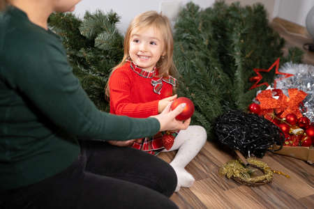 Mother and daughter prepare to decorate the Christmas tree together, sitting on the floor mother gives her daughter red toy ball.の写真素材