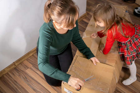 Mother with scissors in her hands with her little girl un packing the cardboard big box with Christmas tree inside.の写真素材