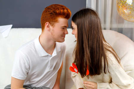 Couple in love brunette girl and red hair boy looking at each other with romantic feelings and girl holding red heart cardboard flowers.の写真素材